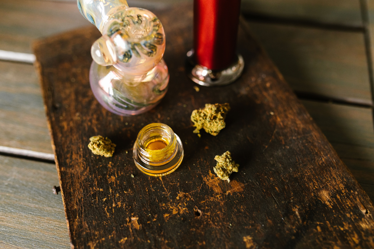 Small jar of cannabis concentrate on a wooden board with cannabis flower and dabbing accessories.