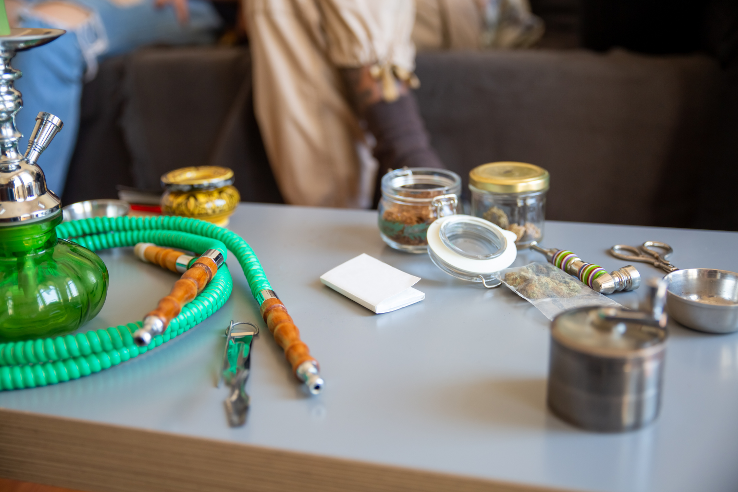 Cannabis smoking devices and accessories arranged on a table, including a glass bong, hose, hand pipes, grinder, jars, and rolling paper