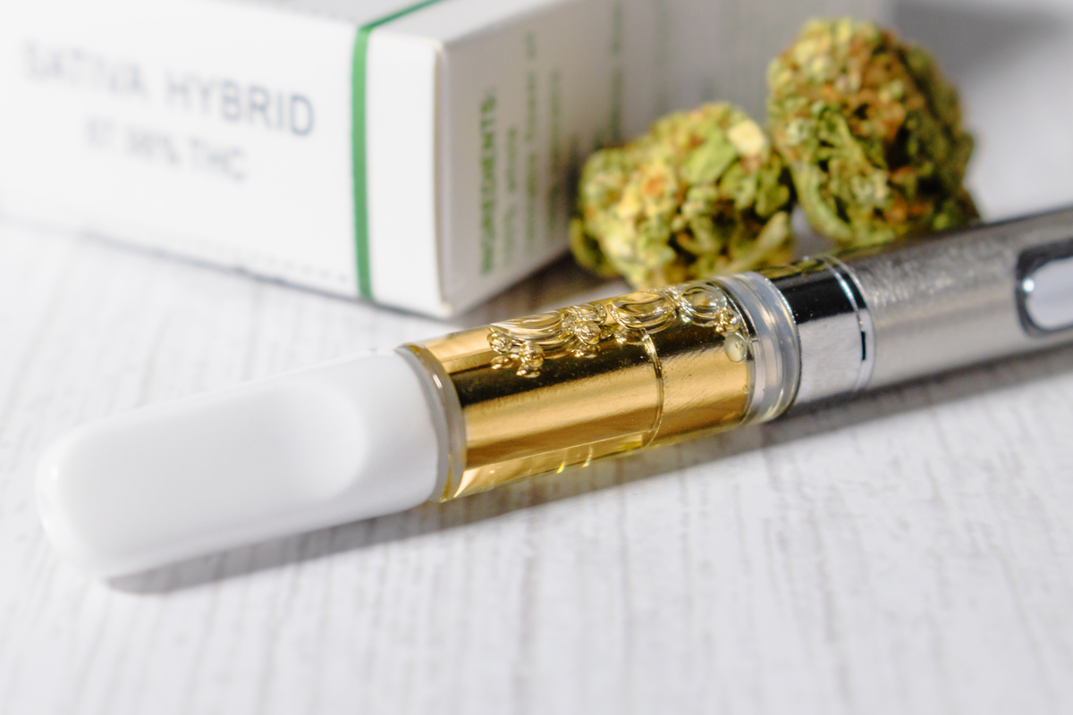 A close-up of a cannabis vape cartridge filled with golden oil attached to a metallic vape pen, with cannabis buds and a product box labeled “SATIVA HYBRID” and “87.39% THC” in the background.