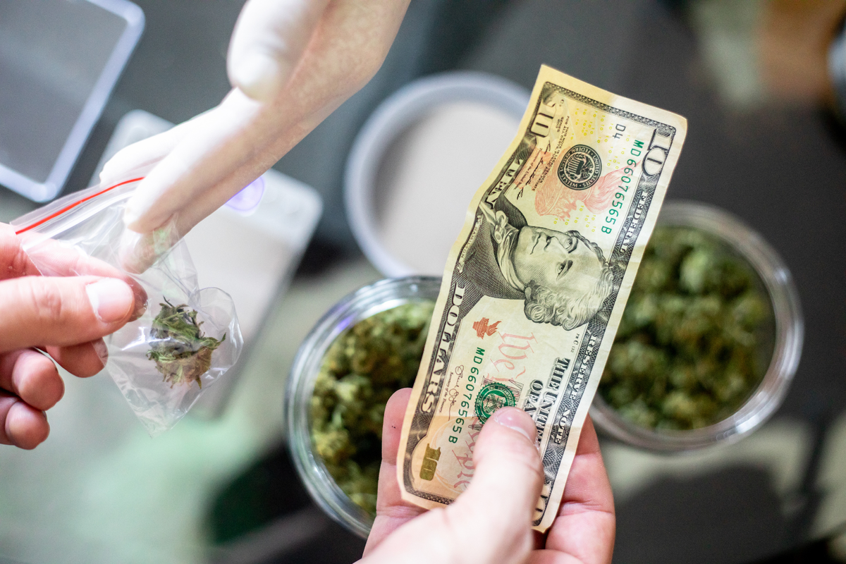 Close-up view of a customer handing a ten-dollar bill to a gloved budtender in exchange for a small plastic baggie of marijuana flower at a counter with glass jars of buds.
