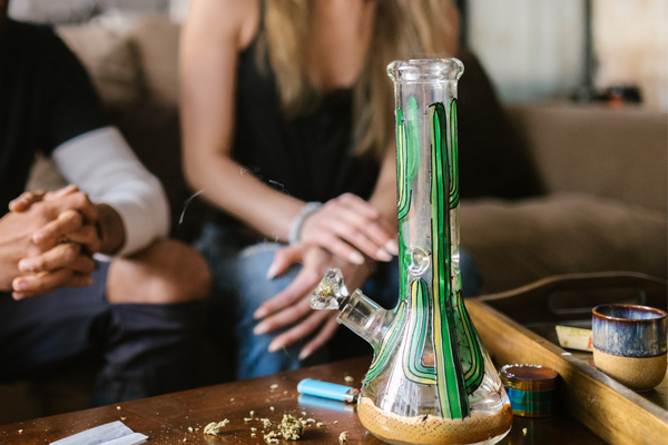 Green glass bong with cannabis accessories and adults seated around a table