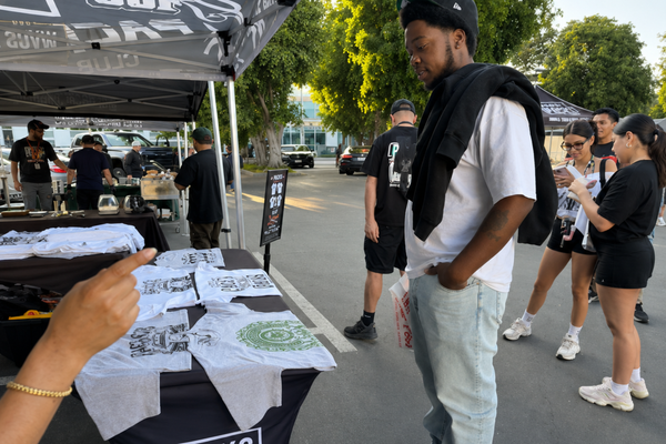 A person points at t-shirts displayed on a table under a tent, with other people browsing in the background.