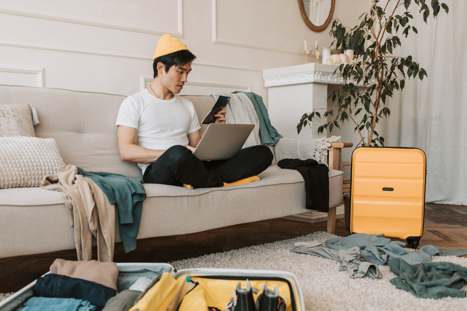Person sitting on a sofa with smartphone and laptop, surrounded by suitcases and clothes while preparing for travel, symbolizing convenience of recreational cannabis delivery during trips.