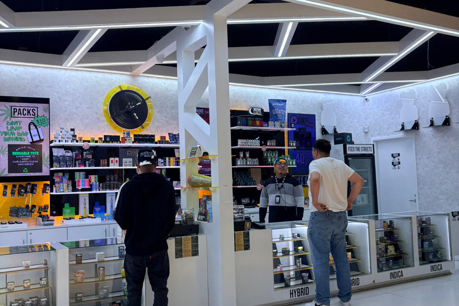 People browsing in a recreational dispensary with glass display cases and shelves of products, a circular yellow-and-black wall sign, and a ceiling of white metal beams.