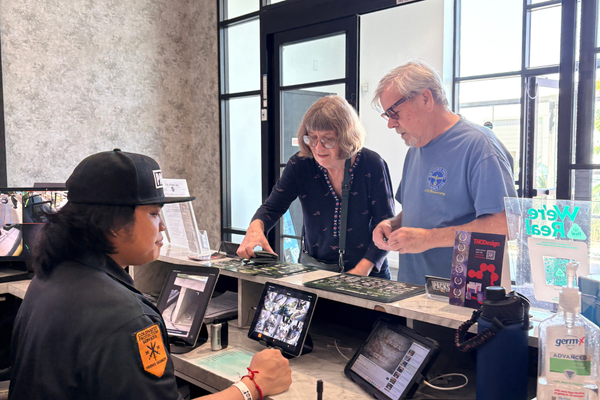 An employee wearing a black cap and uniform with an orange patch assists two customers returning weed at a counter with tablets and displays.